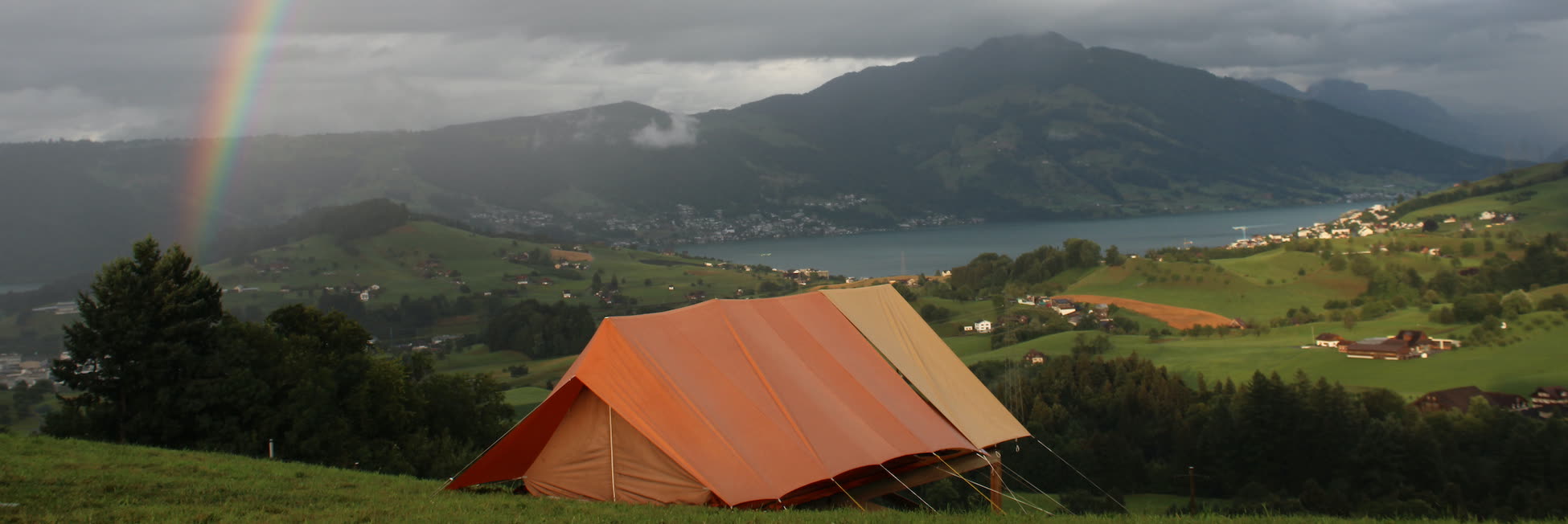 Oranges Zelt auf einer Wiese, darüber ein Regenbogen über dem See